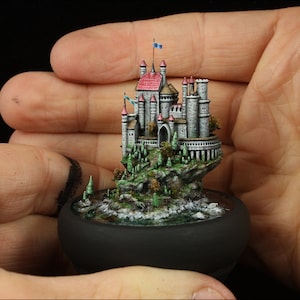 Può includere: Un castello in miniatura con un tetto rosso e muri in pietra grigia si trova su uno sperone roccioso con alberi verdi. Il castello è circondato da un fossato con una piccola cascata. Il castello è tenuto nella mano di una persona.