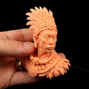 May include: A peach-colored resin bust of a figure wearing an elaborate feathered headdress and a detailed collar featuring a central circular medallion with sun-like carvings and geometric patterns, sculpted with intricate facial features.