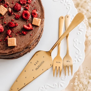 May include: A chocolate cake with raspberries and caramel squares sits on a white, scalloped cake stand. A gold cake server and two forks, engraved with "mr." and "mrs." and the date "08.08.2026", are arranged on the stand.