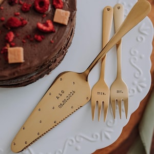 Puede incluir: Un pastel de chocolate cubierto con frambuesas y cuadrados de caramelo sobre un plato blanco decorativo. Servidor de pastel y tenedores dorados con "Mrs." y "Mr." junto al pastel. El servidor está grabado con iniciales y una fecha.