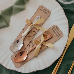 May include: Two metal spoons, one silver and one copper-toned, each tied with a gold ribbon to a rectangular kraft paper card. The silver spoon is engraved with the name Adam and a star, while the copper spoon features the name Eva and a heart.