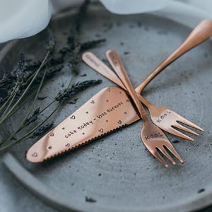 Puede incluir: Un servidor de pastel y dos tenedores de color cobre descansan sobre un plato gris, con ramitas de lavanda. El servidor está inscrito con "cake today - love forever" y diseños de corazón. Los tenedores están inscritos "Kool".
