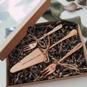 May include: A copper-colored wedding cake serving set in a gift box. The set includes a cake server, two forks, and a spoon, all tied with twine. The cake server has the inscription "love comes in clutch."