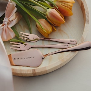 Könnte beinhalten: Ein Set aus drei Servierbesteck aus Roségold auf einem Holztablett mit einem Strauß orangefarbener Tulpen. Das Besteck ist mit den Worten "love is sweet", "Adam" und "Hana" graviert.