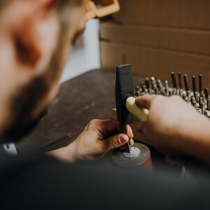 May include: A person uses a hammer to strike a metal object on a red base. The hammer has a yellow handle and a black head. There are many small metal tools in a wooden holder in the background.