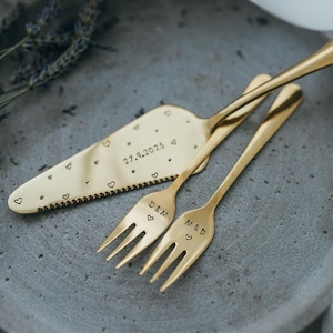 Könnte beinhalten: Goldfarbenes Tortenmesser und zwei Gabeln auf einem grauen Teller. Das Messer ist mit Herzen und dem Datum 27.9.2025 graviert. Die Gabeln sind mit Initialen und Herzen graviert, ideal für eine Hochzeit.