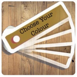 以下が含まれることがあります： 「Choose Your Colour」の文字が入った、金と白のデザインのカラーサンプルファン。ファンは、白い縁取りのある、いくつかの茶色の色合いを表示しています。背景は木製の表面です。