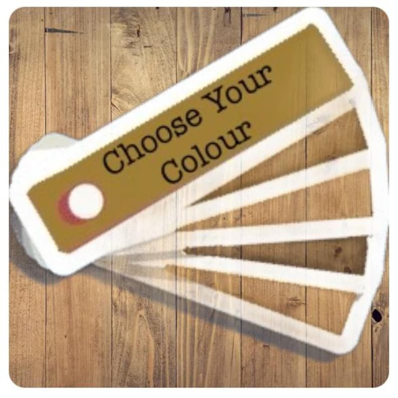 以下が含まれることがあります： 「Choose Your Colour」の文字が入った、金と白のデザインのカラーサンプルファン。ファンは、白い縁取りのある、いくつかの茶色の色合いを表示しています。背景は木製の表面です。