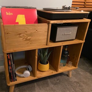 以下が含まれることがあります： 複数のコンパートメントを備えた木製のレコードプレーヤー台。 レコードプレーヤー、スピーカー、ビニールレコード、小さな鉢植えの植物を収納。 赤と黄色のビートルズのアルバムが見えます。 台は明るい色の木材で作られています。