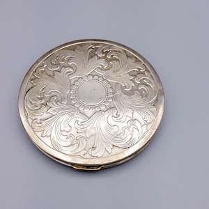 Op de afbeelding: Een ronde, zilveren compact met ingewikkelde bloemen- en bladgravures. De compact heeft een centraal cirkelvormig ontwerp omgeven door een parelrand. Het metaal heeft een gepolijst, reflecterend oppervlak en een kleine gouden sluiting.