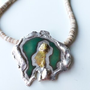May include: A silver necklace with a green and gold pendant. The pendant is shaped like a rock with a rough, irregular edge. The pendant has a green enamel inlay and a gold accent.