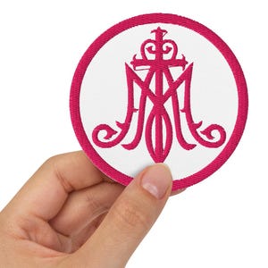 以下が含まれることがあります： 白い背景に鮮やかなピンクの縁取りが施された円形の刺繍ワッペン。中央には鮮やかなピンクのモノグラムシンボルが刺繍されています。デザインは、十字架と王冠が絡み合った様式化された文字「M」で構成されています。