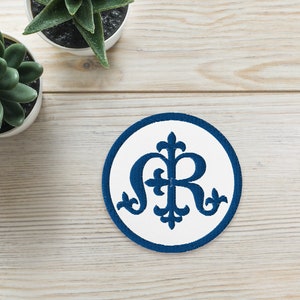 Könnte beinhalten: Ein weißer Kreis mit einem blauen gestickten Design der Initialen der Jungfrau Maria, "M" und "R", mit einem Kreuz in der Mitte. Zwei grüne Sukkulenten in Töpfen stehen auf einer weißen Holzoberfläche.