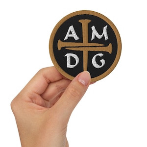 Peut inclure: Un patch rond, marron, avec une croix noire et les lettres "AMDG" en blanc. La croix a quatre clous.