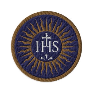 May include: A round, embroidered patch with a brown outer ring and a blue inner circle. The inner circle features a sunburst design with a white cross and the letters "IHS" in the center.