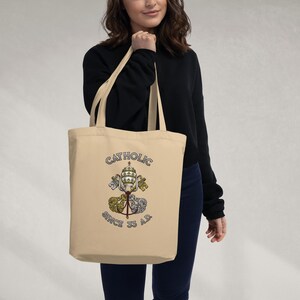 May include: A beige tote bag featuring the word "CATHOLIC" in blue and gold. The bag displays a crest with a crown, keys, and religious symbols, with the text "SINCE 33 A.D." underneath. The bag has long handles.