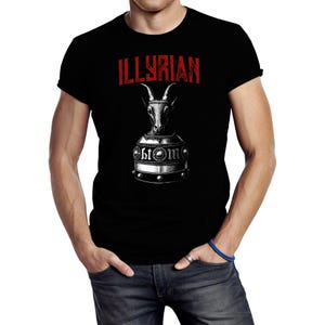 Könnte beinhalten: Schwarzes T-Shirt mit dem Wort "ILLYRIAN" in Rot über einer detaillierten Illustration eines Ziegenkopfes in einem Metallhelm. Der Helm hat die Buchstaben "btom".