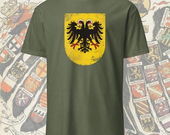 May include: Olive green t-shirt featuring a distressed yellow shield with a black double-headed eagle emblem. The shirt has a classic crew neck and short sleeves. The text "Das hadig romich rech" is visible at the top. The design is reminiscent of heraldry.