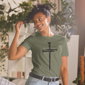 May include: Olive green t-shirt featuring a black cross design made of nails. The short-sleeved shirt has a crew neck and is made of a soft material. The graphic is centered on the front of the shirt.