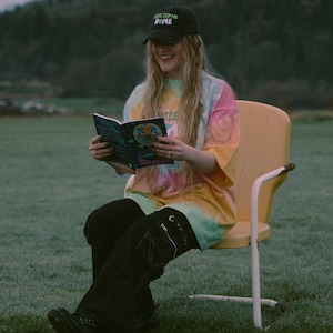 May include: A person wearing a black baseball cap with the text "Music Led Me Home" and a tie-dye t-shirt is reading a book. The person is sitting on a yellow chair in a grassy field. The book has a colorful illustration.