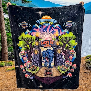 Puede incluir: Manta negra con un diseño psicodélico colorido que presenta un T-Rex, OVNIs y el texto "LOST IN THE LANDS". La manta tiene un fondo de cielo estrellado con setas y árboles vibrantes.