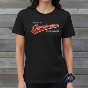 May include: Black t-shirt featuring the text "I GOT LOST IN Copenhagen AND LOVED IT" in red and white. The design includes a red banner. The shirt has a crew neck and short sleeves.