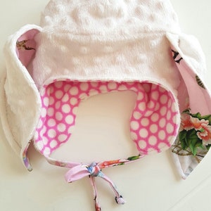 May include: A white, plush baby hat with bunny ear flaps. The interior is pink with white polka dots. The edges of the ear flaps have a floral pattern. A matching floral tie secures the hat.