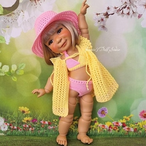 May include: A doll wearing a pink crocheted sun hat, bikini, and yellow crocheted shawl. The doll is standing on a grassy surface with colorful flowers in the background. The doll is smiling and has its arm raised.