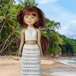 Puede incluir: Muñeca con top de ganchillo blanco y falda a juego. La muñeca tiene cabello castaño y está en una playa con palmeras y cielo azul. El texto "Janette's Doll Studio" es visible.