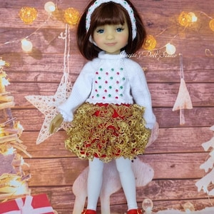Puede incluir: Una muñeca vestida con un atuendo festivo. La muñeca lleva un suéter blanco con detalles rojos y verdes, una falda dorada y roja, medias blancas y zapatos rojos. Una diadema blanca con detalles rojos y verdes adorna el cabello castaño de la muñeca. El fondo tiene decoraciones navideñas.