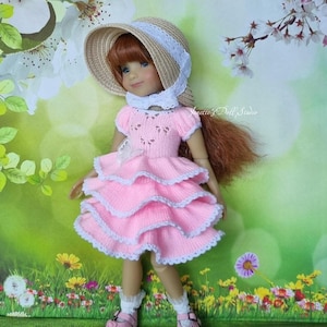 Puede incluir: Una muñeca con un vestido rosa escalonado con ribete blanco, un sombrero a juego y zapatos rosas. La muñeca tiene el pelo rojo y está sobre un fondo verde y floral. El vestido tiene un lazo blanco.