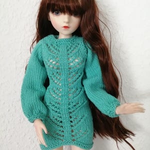 May include: A doll wearing a teal green knitted dress with long sleeves and a decorative openwork pattern down the front. The doll has long brown hair and is wearing black stockings.