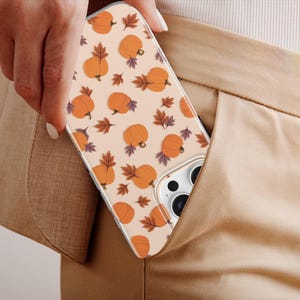 May include: A clear phone case featuring a repeating pattern of orange pumpkins and brown autumn leaves on a light beige background. The case has a cutout for a multi-lens camera system and is shown partially inserted into a tan pocket.