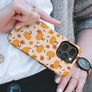 May include: A protective phone case featuring a repeating pattern of orange pumpkins and brown autumn leaves on a light beige background. The case has a precise cutout for the rear camera module, which contains three circular lenses and a flash.