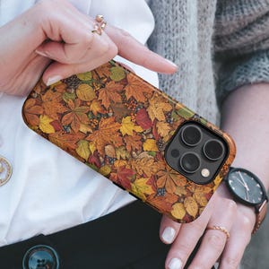 May include: A rectangular smartphone case featuring a detailed autumnal pattern of fallen leaves in shades of orange, yellow, red, and brown, interspersed with small acorns and pinecones, with a black camera lens cutout in the upper corner.