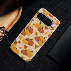 May include: A smartphone case with a light beige background featuring a repeating pattern of orange and yellow pumpkins with green stems and small brown autumn leaves scattered throughout. The case has a rectangular camera cutout at the top.