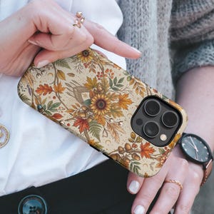 May include: A smartphone case featuring a detailed autumn-themed pattern with sunflowers, orange and yellow maple leaves, acorns, berries, and small crescent moon symbols on a cream-colored background, designed for a device with a triple-lens camera.