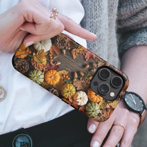 May include: A smartphone case featuring a detailed autumn-themed print of orange and white pumpkins, brown pinecones, acorns, and fallen leaves scattered across a dark wooden surface. The case has a black camera cutout for a triple-lens system.