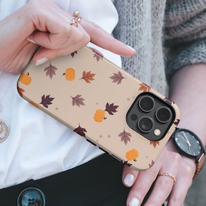 May include: A beige smartphone case featuring a repeating pattern of orange pumpkins and brown autumn leaves. The case has a cutout for a triple-lens camera system on the back, with a black camera housing and a smooth, matte-finish surface design.