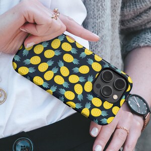 May include: A black smartphone case with a repeating pattern of yellow pineapples and green leaves. The case has a glossy finish and a camera cutout. The phone case is held by a person.