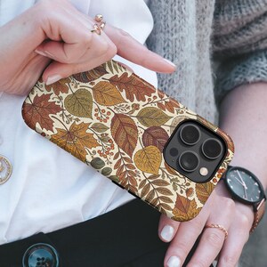 May include: A smartphone case featuring a detailed pattern of autumn leaves in shades of orange, yellow, brown, and green. The design includes intricate line work on the foliage, small berries, and twigs against a cream-colored background.