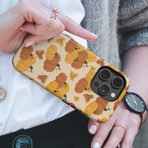 May include: A smartphone case featuring a repeating pattern of orange and yellow pumpkins with brown stems and small brown autumn leaves on a light beige background. The case has a black camera module cutout and a black border around the lens area.