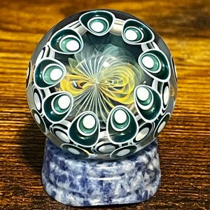 May include: A clear glass paperweight with a swirling yellow and white design at its core, surrounded by a ring of green and white teardrop shapes. It rests on a blue and white speckled stone base, set against a wooden surface.