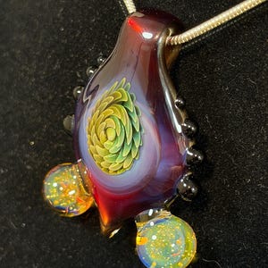 May include: A handcrafted glass pendant with a spiral design in shades of green and yellow, encased in a teardrop-shaped glass. The pendant has a deep red and purple hue, with two small, round, colorful glass accents. It hangs on a silver chain.
