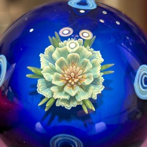 May include: A detailed glass art sphere with a vibrant blue background. The sphere features a central, light green and yellow flower design with intricate petal details. Green leaves and white swirl accents complete the design.