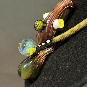 May include: A handcrafted glass pipe with a swirling galaxy design in blue, yellow, and green. The pipe features a brown and gold stem with yellow and white accents. A gold-colored chain is visible.