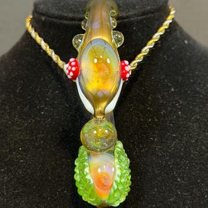 May include: A colorful glass pendant necklace with a gold chain. The pendant features a yellow and orange center with red mushroom accents and a green base. The glass has a textured, iridescent finish.