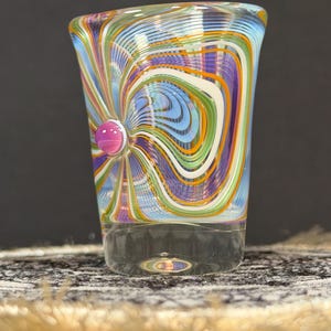 May include: A vibrant, hand-blown glass tumbler featuring a swirling design in blue, green, orange, purple, and white. A small, pink glass bead is fixed to the side. The lower part of the glass is transparent.