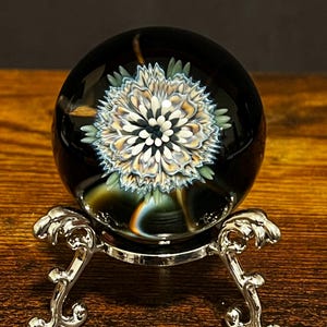 May include: A black glass orb featuring a detailed floral pattern in white, blue, and tan. The orb is presented on a decorative silver metal stand with ornate, curved legs. The background is a dark brown.