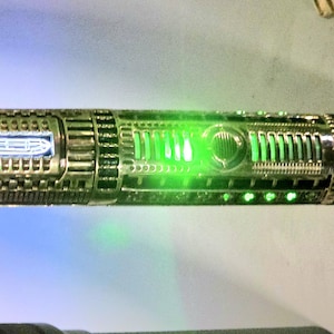 Puede incluir: Un detallado sable de luz con acabado dorado y plateado. La sección central brilla en verde brillante, con un panel azul iluminado cerca de la empuñadura. El diseño presenta intrincados detalles mecánicos.